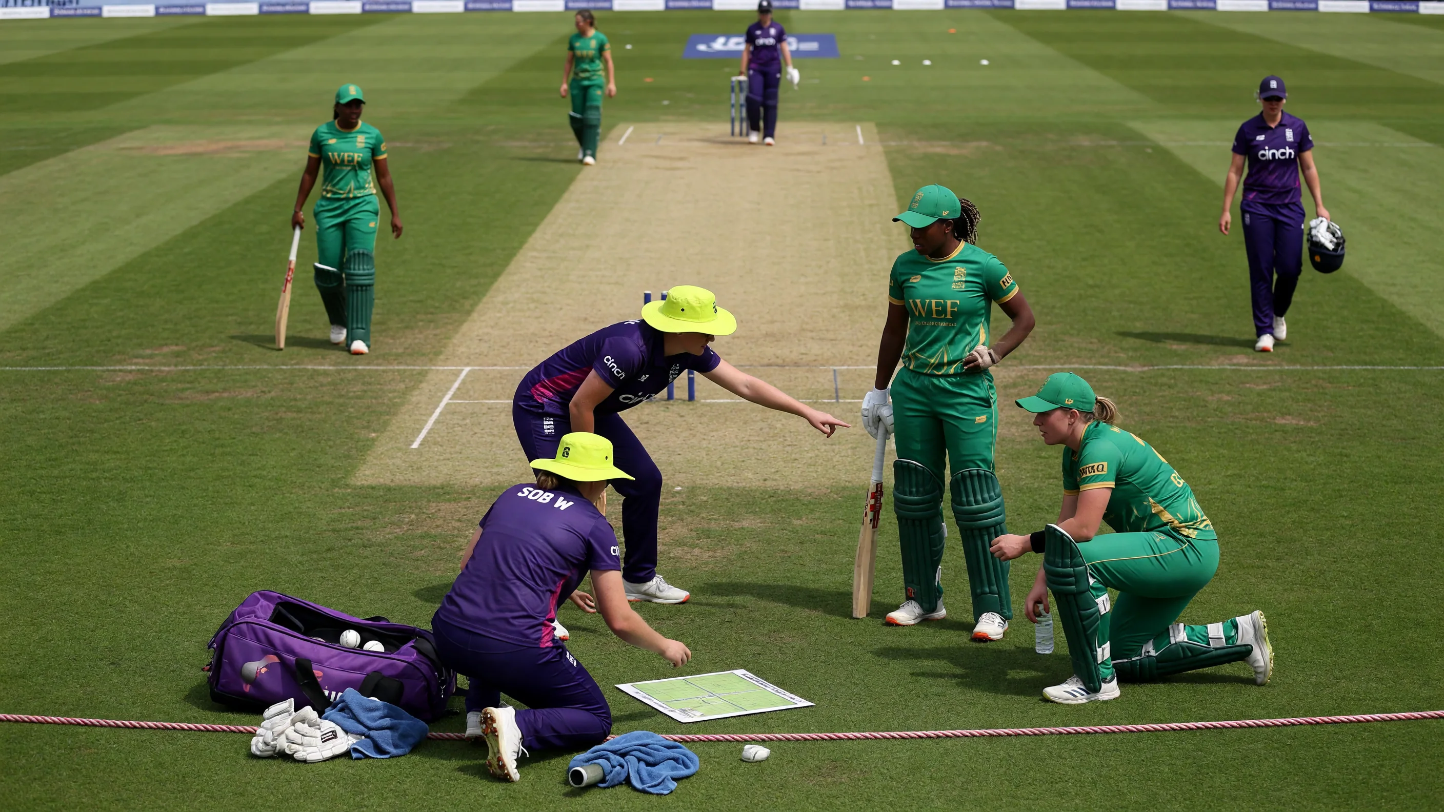 Medium tactical view of fielders setting positions on a sunlit outfield with stumps and players in match kits
